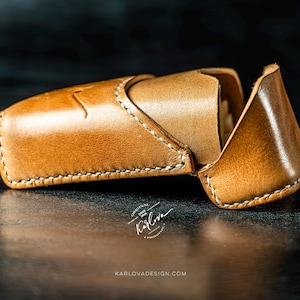May include: A tan leather case with a flap closure and white stitching. The case features a debossed logo and the text Karlova Original Since 2020, with the website URL KARLOVADESIGN.COM printed below the object on the dark surface.