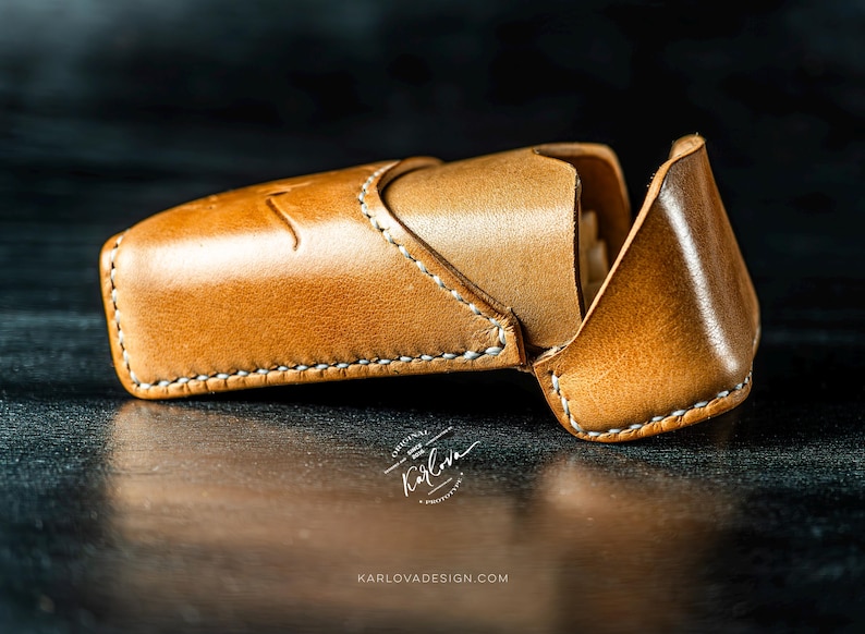 May include: A tan leather case with a flap closure and white stitching. The case features a debossed logo and the text Karlova Original Since 2020, with the website URL KARLOVADESIGN.COM printed below the object on the dark surface.
