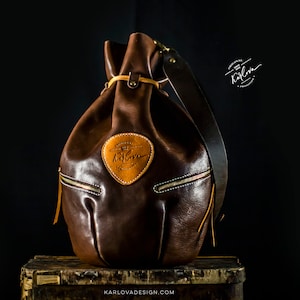 Könnte beinhalten: Eine braune Ledereimer-Tasche mit einem beigefarbenen Lederriemen und einem beigefarbenen Leder-Patch mit dem Text "KARLOVA DESIGN". Die Tasche steht auf einer Holzoberfläche.