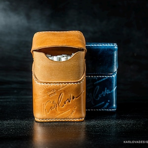 May include: A tan leather cigarette case with white stitching and the embossed text Karlova, positioned in front of a dark blue leather cigarette case with matching white stitching and embossed branding, both featuring a flap closure design.