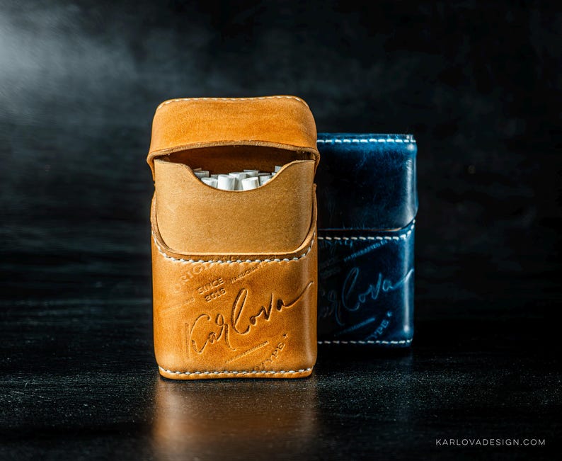 May include: A tan leather cigarette case with white stitching and the embossed text Karlova, positioned in front of a dark blue leather cigarette case with matching white stitching and embossed branding, both featuring a flap closure design.