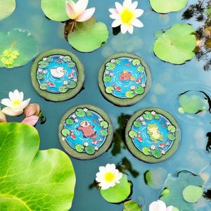 May include: Four round coasters with pond scenes featuring a duck, a frog, an otter, and a fish. Each coaster has a blue centre with lily pads, koi fish, and a cartoon animal. The coasters are surrounded by a green border. The image includes water lilies and flowers.