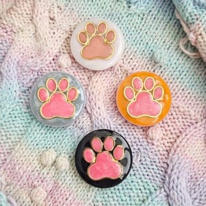May include: Four round, colorful paw print designs with gold trim. The paw prints are pink and set against backgrounds of white, gray, orange, and black. The items are displayed on a pastel-colored knitted fabric.