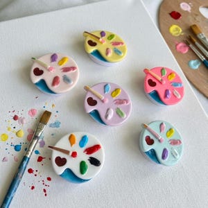 May include: A collection of colorful, miniature artist palette-shaped accessories. Each palette features a heart, a paintbrush, and dabs of paint in various colors. The accessories are set on a white surface, with paint splatters and paintbrushes nearby.