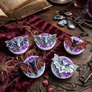 May include: Five circular phone grips featuring sculpted bat figures in various colours including silver, purple, and red. Each bat is mounted on a white base with purple glitter accents. The bats have detailed wings and bodies, displayed as a collection.