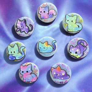 May include: A collection of eight round, colorful cat-themed buttons. Each button features a unique cartoon cat design in various colors like purple, blue, pink, and green, with glitter accents. The background is a gradient of purple and blue.