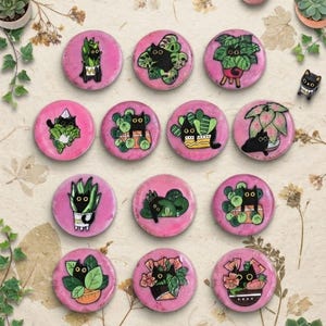 May include: A collection of eleven circular pink pins featuring illustrations of black cats nestled among various green houseplants. Each pin displays a unique cat and plant combination, showcasing detailed line work and vibrant colors on a round surface.