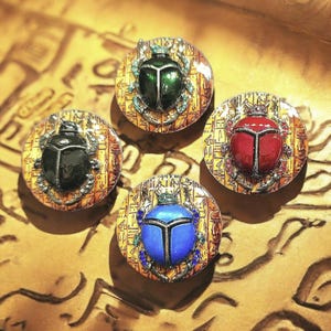 May include: Four round, decorative scarab beetle pendants in green, black, red, and blue. Each beetle is set on a gold-toned background with hieroglyphic designs. The beetles have silver-colored metal accents and are embellished with small gemstones.