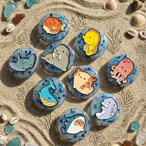 May include: A collection of round, ocean-themed resin art pieces featuring various sea creatures like a shark, whale, and octopus. The pieces are set on a sandy surface with seashells and sea glass.