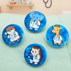 May include: Four circular pins featuring cartoon animals dressed as medical professionals. The designs include a blue elephant, a brown otter with a magnifying glass, a yellow giraffe, and a brown hedgehog holding a blue clipboard, all on blue backgrounds.