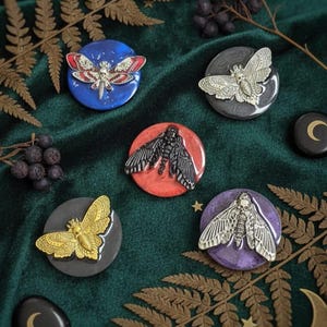 May include: A collection of decorative moth pins with intricate details. The pins feature various colors and designs, including gold, silver, and black moths on round bases. The background is a dark green fabric with fern and berry accents.