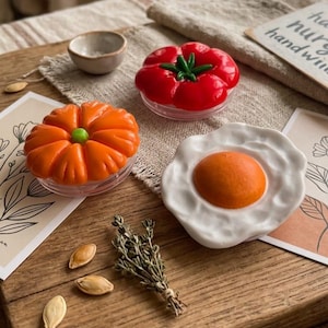 May include: A collection of decorative food-themed items, including a bright orange pumpkin, a red tomato, and a fried egg, all resting on clear, round bases. The items are arranged on a wooden surface with a small bowl and sprigs of herbs.