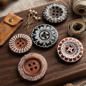 May include: Five round wooden buttons with four holes each, featuring intricate white painted patterns. The buttons vary in color from light brown to dark brown and are arranged on a dark wooden surface alongside sewing pins and a tape measure.
