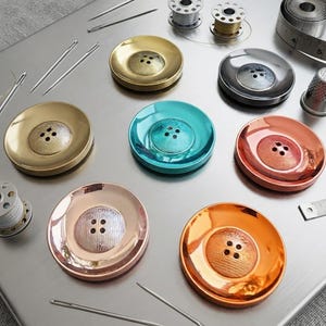 May include: A collection of round, four-hole buttons in metallic finishes including gold, turquoise, copper, silver, and bronze, arranged with sewing needles, metal bobbins, a thimble, and a measuring tape on a smooth, brushed metal surface.