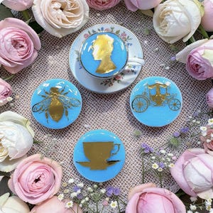 May include: A light blue teacup and saucer set with gold accents, surrounded by four matching coasters. The coasters feature gold silhouettes of a profile, bee, carriage, and teacup. The scene is framed by pink and white roses.