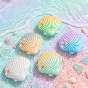 May include: Five shell-shaped phone grips arranged on a sandy surface. Each grip features a gradient color finish, small white faux pearls, and a tiny gold star accent. The colors include white, yellow-green, peach, and purple-blue gradients.