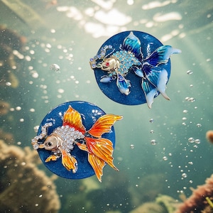 May include: Two bejeweled goldfish brooches. One has orange and yellow fins, the other has blue fins. Both are set on a dark blue circular base. The brooches are detailed with rhinestones and appear to be floating in water.