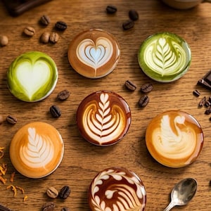 May include: Seven circular coasters featuring detailed latte art designs, including hearts, rosettes, and a swan. The coasters display various shades of brown, tan, and green, mimicking coffee and matcha beverages, arranged on a wooden surface.