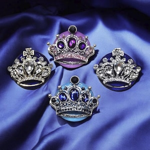 May include: Four bejeweled crown brooches in shades of blue, purple, and silver. Each crown features intricate detailing with sparkling gemstones and a metallic silver frame. The brooches are set against a deep blue satin background.