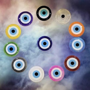 May include: A collection of twelve colorful, round, evil eye charms. The charms feature a blue iris, white and light blue accents, and a black pupil. The charms are in various colors, including yellow, green, blue, orange, and pink.