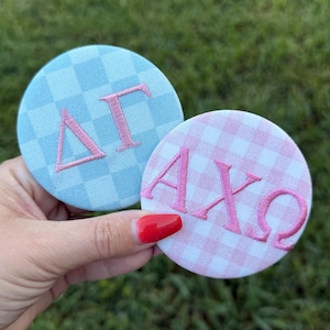 Può includere: Due toppe circolari in tessuto con lettere greche ricamate. Una toppa presenta un motivo a scacchi azzurro e bianco con lettere Delta Gamma rosa. L'altra ha un motivo a quadretti rosa chiaro e bianco con lettere Alpha Chi Omega rosa.