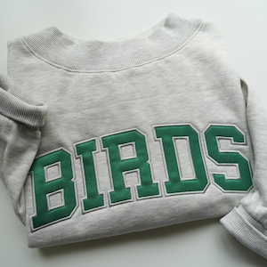 Könnte beinhalten: Ein hellgrau meliertes Langarm-Sweatshirt mit dem Wort BIRDS in großen, fetten, waldgrünen Blockbuchstaben mit weißer Umrandung. Das Kleidungsstück verfügt über einen gerippten Rundhalsausschnitt und gerippte Ärmelbündchen im klassischen, sportlichen Design.