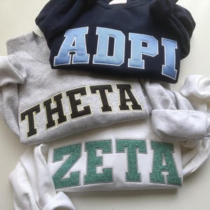 Op de afbeelding: Drie sweaters met ronde hals op elkaar gestapeld. De bovenste is marineblauw met lichtblauwe ADPI-letters. De middelste is grijs gemêleerd met zwarte THETA-letters. De onderste is wit met groene glitter ZETA-letters in een college-lettertype.