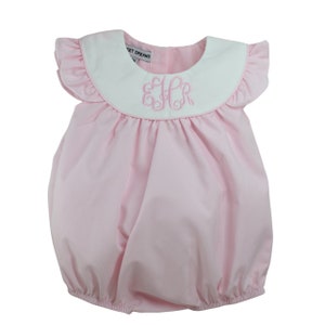 May include: Pink baby bubble romper with a white ruffled collar and sleeves. The collar features an embroidered monogram. The romper has the text "SWEET DREAMS" on the back of the collar.