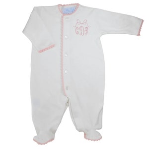 May include: White baby onesie with pink crocheted trim and a pink embroidered monogram with a bow. The monogram reads "CDR".