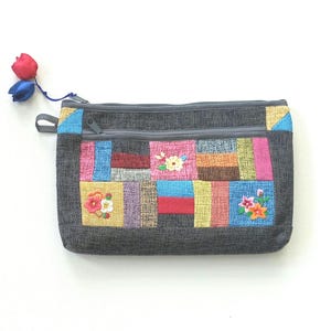 May include: A grey zippered pouch with a patchwork design in various colours and floral embroidery. A small red and blue flower charm is attached to the zipper pull. The pouch is rectangular and appears to be made of fabric.
