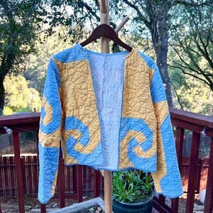 May include: A blue and yellow quilted jacket with a unique pattern. The jacket is hanging on a wooden hanger.
