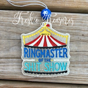 May include: A novelty air freshener with a circus tent design in red and white stripes, with the text "RINGMASTER OF THE SHIT SHOW" in blue and light blue. It has a blue star-studded bead and a white string for hanging.