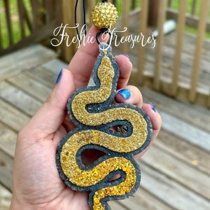 May include: A glittery gold and black snake-shaped air freshener with a black string and a gold ball at the top. The text "Freshie Treasures" is written in cursive. The air freshener is held in a hand.