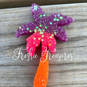 May include: A pink, orange, and purple glitter palm tree shaped air freshener. The palm tree is on a wooden surface.