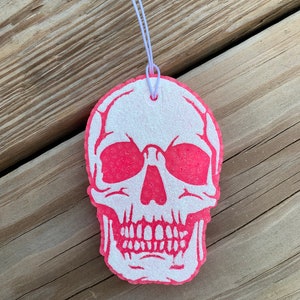 May include: A white skull with a pink glitter background. The skull is hanging from a white string.