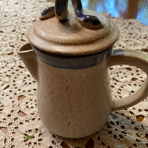 May include: Tan ceramic teapot with a dark blue accent band and handle. The lid features a decorative, curved handle. The teapot has a textured surface and a spout for pouring. The teapot is sitting on a cream-colored lace tablecloth.
