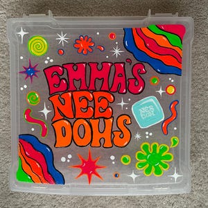 May include: A clear plastic storage case featuring hand-painted, colorful designs including rainbow waves, stars, and dots. The text "EMMA'S NEE DOHS" is written in large, bold, orange and red letters, with a small light blue square icon labeled "NEE DOH".
