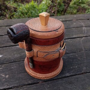 Könnte beinhalten: Eine Holztöpfchen für Tabak mit einem Lederriemen und einem goldfarbenen Deckel. Eine Pfeife liegt an der Seite des Töpfchens.