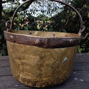 Pode incluir: Uma panela de cobre antiga grande com uma alça de metal. A panela tem uma pátina desgastada e envelhecida e é decorada com rebites ao redor da borda.