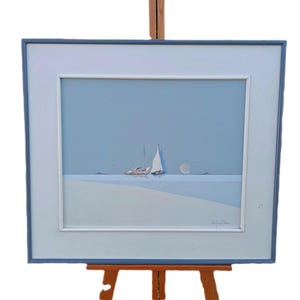 Puede incluir: Pintura enmarcada de un paisaje marino con dos veleros sobre el agua bajo un cielo azul claro. La pintura tiene un borde blanco y un marco azul. La obra de arte se exhibe en un caballete de madera.