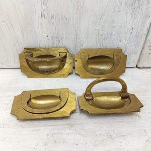 May include: Four antique brass drawer pulls with a rounded rectangular shape and a recessed handle. The pulls have a slightly ornate design around the edges.