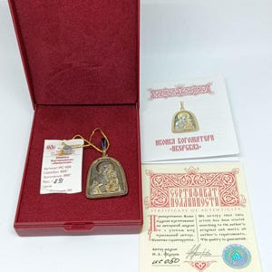 May include: A religious pendant in a red box. The pendant is silver and gold-toned, featuring an iconographic design. Accompanying documents include a certificate of authenticity and a card with text in a foreign language. The box is open, revealing the pendant and documents.