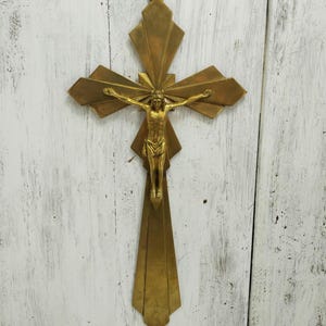 Puede incluir: Un crucifijo dorado con una figura detallada de Jesucristo. La cruz tiene un diseño geométrico con líneas radiales. La figura está en posición de crucifixión, con los brazos extendidos. El crucifijo está colgado sobre una superficie de madera blanca.