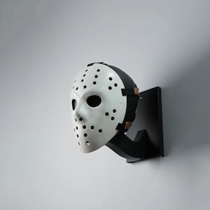 May include: A white hockey-style mask with numerous circular ventilation holes and black eye openings, mounted on a black wall-hanging display stand. The mask features a black strap system with metallic fasteners securing it to the display mount.