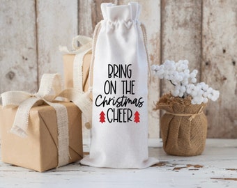 以下が含まれることがあります： 黒い文字で「Bring on the Christmas Cheer」と書かれた、赤いクリスマスツリーのアイコンが2つ付いた白い布製のワインボトルバッグです。