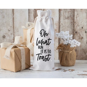 Peut inclure: Sac en tissu blanc pour bouteille de vin avec du texte noir qui dit "Oh what fun it is to toast" avec des accents de flocons de neige blancs.