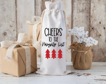 以下が含まれることがあります： 黒い文字で「Cheers to the Naughty List」と書かれた白い布製のワインボトルバッグと、赤いクリスマスツリーが3本。