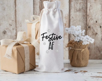 以下が含まれることがあります： 黒い文字で「Festive AF」と雪の結晶のデザインが施された白い布製のワインボトルバッグです。