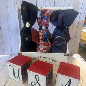 May include: A white wooden crate holds three decorative wine bottle covers. The covers are navy blue, and American flag-themed. Three red, white, and blue wooden blocks spell out "USA". The crate has the text "LIQUES & CRISTAUX DE BOHEME".