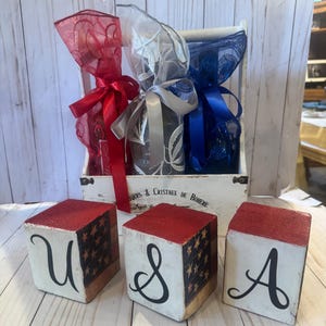 May include: A white wooden crate with three bottles wrapped in red, silver, and blue decorative mesh and ribbons. Three wooden blocks spell out "USA" with a red top, a painted American flag design, and black lettering.